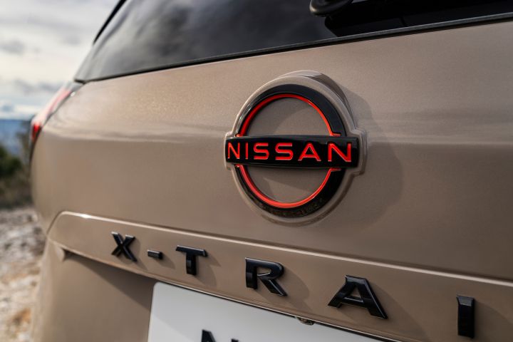 Nahaufnahme des neuen 2026 Nissan X‑Trail mit Nissan-Logo und X‑Trail-Emblem auf der Rückseite des beigen Fahrzeugs. Das Nissan-Logo besteht aus einem magmaroten, beleuchteten Schriftzug innerhalb eines kreisförmigen Emblems, wodurch die Markenidentität und die Modellbezeichnung hervorgehoben werden.