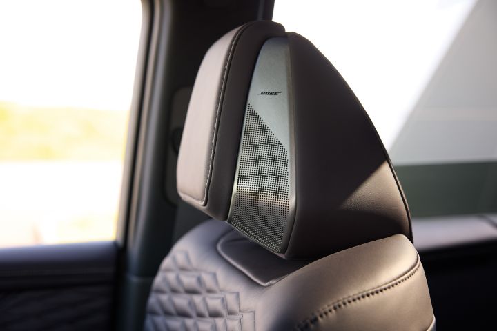 新型エルグランドのシートヘッドレストをクローズアップした画像で、右方向からの斜めの視点で捉えられている。ブラックのレザー調ヘッドレスト側面には、パンチング加工が施されたシルバーカラーのスピーカーグリルが組み込まれており、「BOSE」のロゴが刻印されている。シート背面にはダイヤモンドステッチのデザインが見え、質感の高い素材感が強調されている。背景にはサイドウインドウから差し込む明るい自然光があり、室内の一部が柔らかくぼかされている。