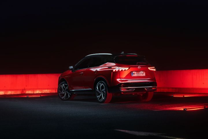 Ansicht von schräg hinten auf einen Nissan Qashqai in Fuji Sunset Red im Dunkeln auf einem Parkplatz mit roten Leuchtakzenten vor dem schwarzen Hintergrund