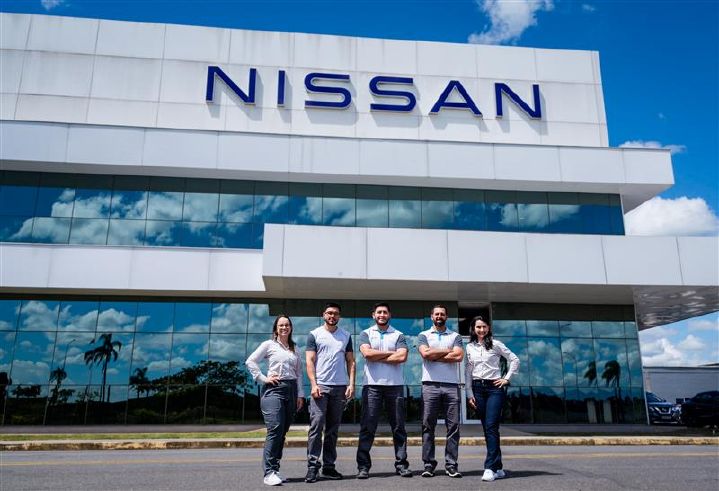 Cinco funcionários da Nissan,de uniforme, estão de pé na frente do prédio administrativo da fábrica, um prédio espelhado com logo da Nissan no alto