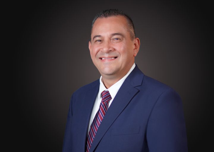Ricardo smiling in a blue suit against a gray background.