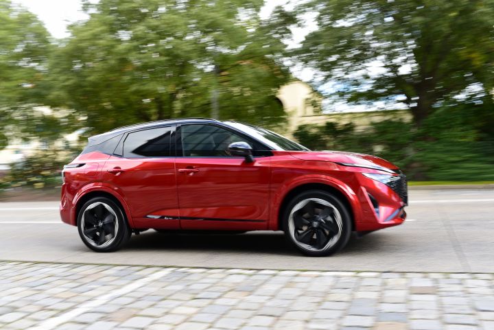 Dynamische seitliche Aufnahme eines roten Nissan Qashqai, der über eine belebte Straße fährt, Auto und Hintergrund sind leicht verschwommen