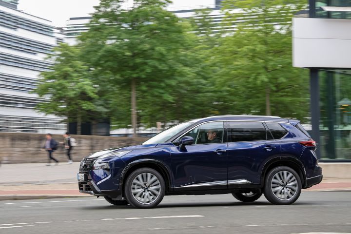 Ein dunkelblauer Nissan X‑Trail fährt seitlich an einem städtischen Platz vorbei