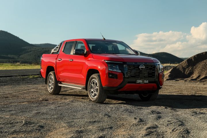 Front three-quarter view of a 2026 Nissan Navara ST-X dual cab in Outback Red, parked on gravel with low hills, open sky, and a dark earth mound behind the vehicle.