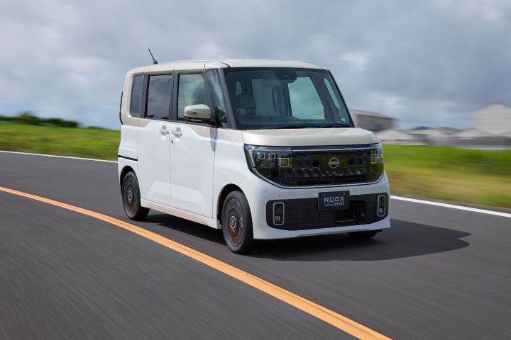 The new Nissan Roox kei car, featuring a white pearl / frozen vanilla pearl two-tone, is driving on a road with a yellow center line. The background includes green grass and buildings under a cloudy sky.