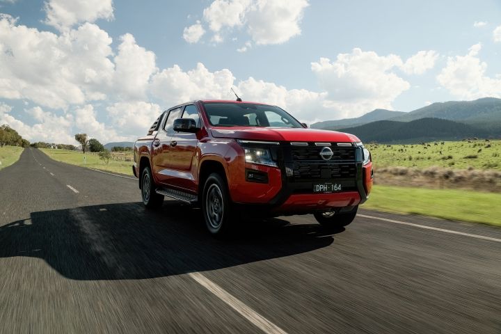 Front three‑quarter view of a red 2026 Nissan Navara ST‑X driving on an open rural road flanked by grassy paddocks and distant hills.