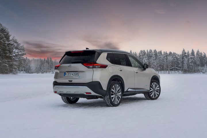 Silberner Nissan X‑Trail e‑Power e‑4ORcE steht auf schneebedecktem Eissee mit verschneiten Bäumen und Holzhütten im Hintergrund, Abendhimmel, 3/4 Heck