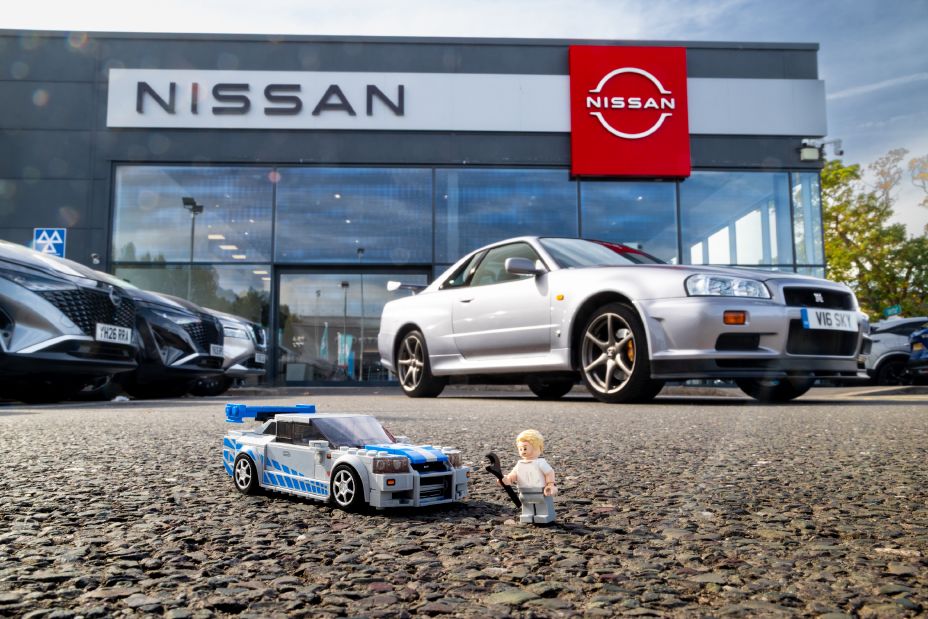 Blue and silver LEGO Nissan Skyline GT‑R model and LEGO minifigure displayed outside a Nissan dealership with a silver Nissan Skyline GT‑R in the background.