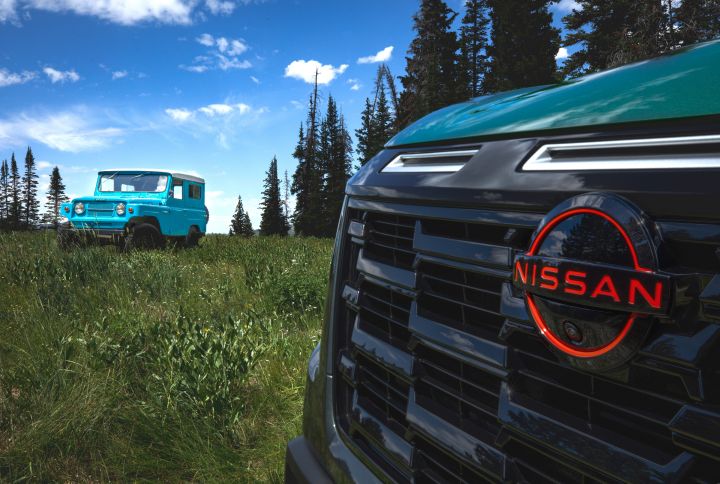 Blue 1967 Nissan Patrol in a piney meadow with boue skies behind, a Nissan SUV's grill is parked close to the camera shot.