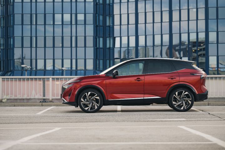 Seitenansicht eines Nissan Qashqai in Fuji Sunset Red auf einem Parkplatz vor einem verglasten Hotelgebäude