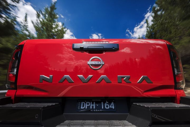 Low-angle rear detail of the tailgate on a 2026 Nissan Navara ST‑X Outback Red dual cab, showing NAVARA lettering, the Nissan badge, and the rear camera with trees and sky behind it.