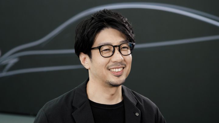 Porträt des Nissan Designers Nobutaka Tase, der in die Kamera lächelt und schwarze Kleidung sowie eine schwarze Brille trägt, im Hintergrund sieht man eine dunkle Wand mit einer Designskizze