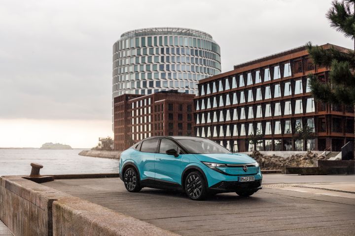 Nissan LEAF kjører langs vannet med moderne bygg i bakgrunnen.