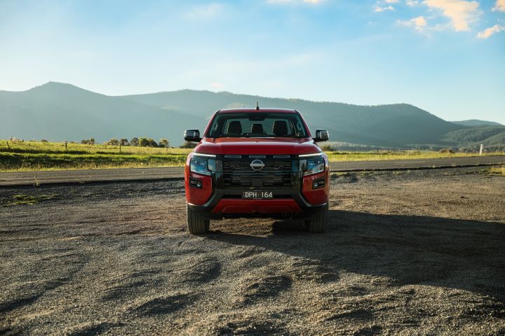 Front view of a 2026 Nissan Navara ST‑X dual cab in Outback Red, parked on gravel with open paddocks and layered hills behind the vehicle.
