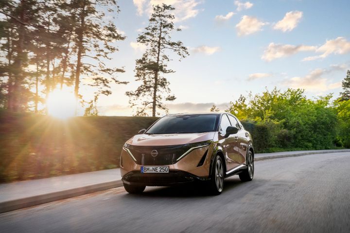 Een Nissan ARIYA die op een weg rijdt met daarachter een ondergaande zon. De Nissan SUV is bronskleurige.