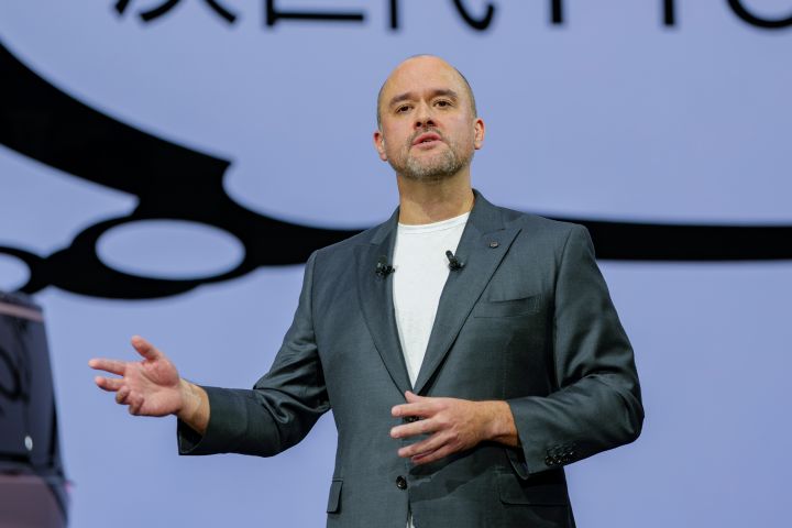 On the stage, CEO Ivan Espinosa, dressed in a suit, stands with his right arm extended while delivering a presentation. In the background, a large screen is visible, and on the left side of the foreground, part of the all-new Elgrand can be seen.