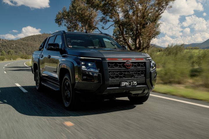2026 Nissan Navara PRO-4X finished in Boulder Grey pictured from a dynamic front-left rolling angle with black mesh grille, red accents and blurred hills.