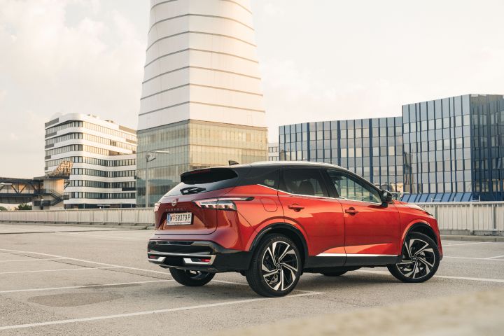 3/4 Heckansicht eines Nissan Qashqai in Fuji Sunset Red vor einem hohen weißen Turm und mehreren verglasten Hochhäusern