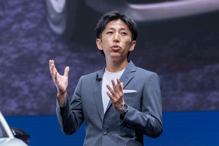 発表会場のステージで新型エルグランドについて説明する中村CPSの上半身を捉えた写真。中村CPSはグレーのジャケットと白いシャツを着用し、正面やや左方向を向きながら両手を上げて説明している。背景には大型スクリーンが広がり、新型エルグランドの車両画像の一部が大きく映し出されている。画面左下にはホワイトの新型エルグランドのフロント部分がわずかに見え、青とグレーを基調とした会場照明がステージを照らしている。新型エルグランドのデザインや技術についてプレゼンテーションを行う様子を捉えた写真。