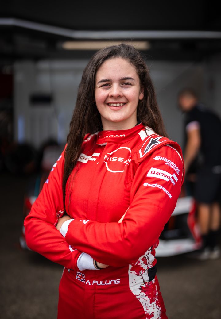 Abbi Pulling, Rookie und Simulator-Fahrerin des Nissan Formula E Teams, steht vor der Teamgarage in ihrem roten Nissan Formula E Team-Rennanzug und lächelt mit verschränkten Armen.