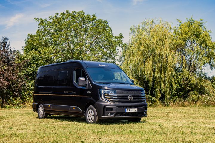 Blick von schräg vorne auf einen schwarzen Nissan Interstar Eifelland Camper, der auf einer Wiese parkt.