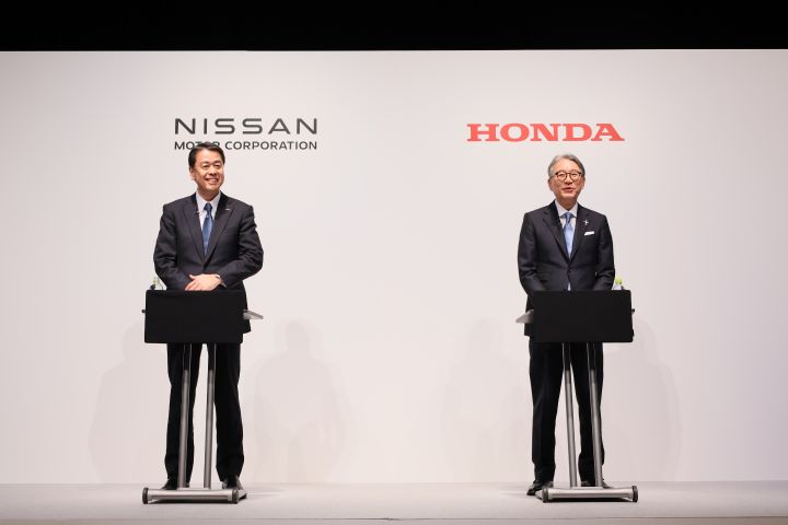 Dois executivos asiáticos de terno, da Nissan e da Honda, estão em cima do palco sorrindo. Acima de cada um deles está o logotipo  da empresa que representam (Nissan e Honda)