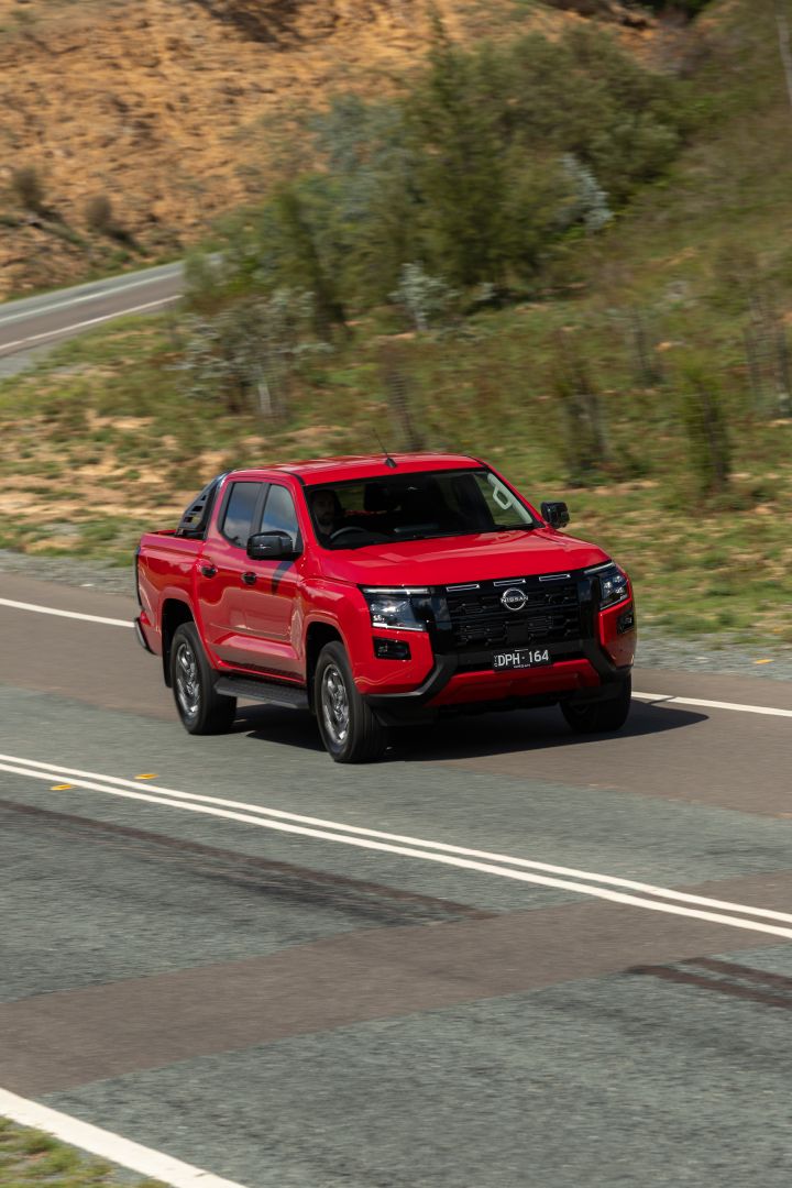 A red 2026 Nissan Navara ST‑X dual cab driving on a curved road in a hilly, semi‑rural landscape.