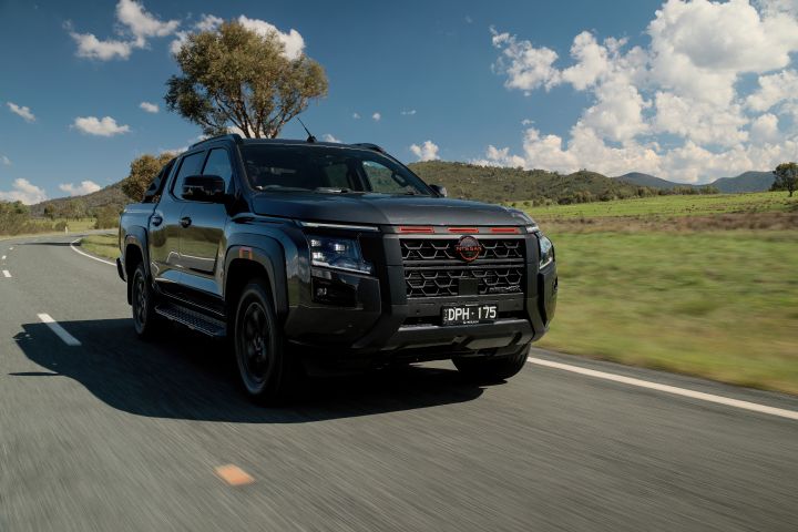 2026 Nissan Navara PRO-4X finished in Boulder Grey shown in a broader front-side tracking shot through open pasture beneath bright clouds and distant ridgelines.