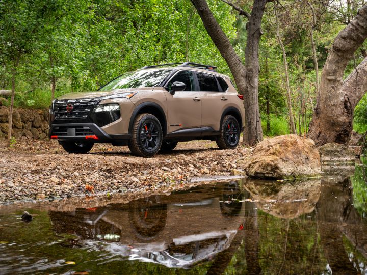 Lower front left 3/4 view of the 2025 Nissan Rogue rock creek parked in front of a wooded creek that is reflecting its image.
