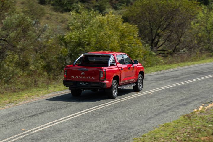 Rear three‑quarter view of a 2026 Nissan Navara ST‑X dual cab finished in Outback Red, travelling along a gently curving rural road bordered by mixed vegetation.