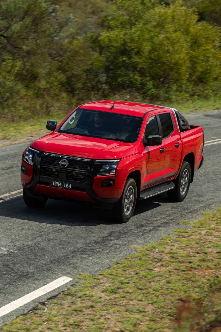 Front three‑quarter view of a 2026 Nissan Navara ST‑X Outback Red dual cab driving on a sunlit country road with rolling hills and cloud patches in the background.