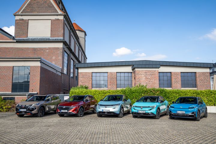 Vor einem Backsteingebäude und einer grünen Hecke stehen nebeneinander die fünf Nissan Modelle Nissan X‑Trail N-Trek, Nissan Qashqai, Nissan Ariya, Nissan Leaf und Nissan Micra, Blick von vorne auf die Gruppe