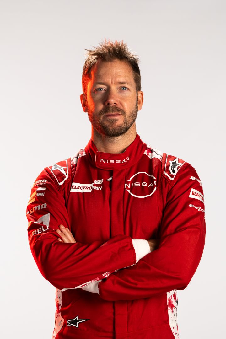 Sam Bird im roten Rennanzug des Nissan Formula E Teams mit vor der Brust verschränkten Armen vor weißem Hintergrund.