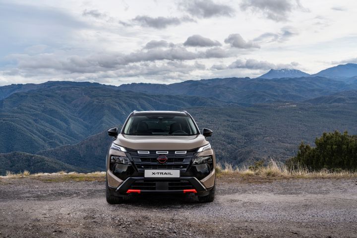 Nissan X‑Trail N-Trek in Baja Storm, geparkt auf einer Schotterstraße mit bergigem Gelände und bewölktem Himmel im Hintergrund. Das Fahrzeug verfügt über eine dunkle Außenlackierung mit magmaroten Akzenten an der vorderen Stoßstange und einem markanten Nissan-Logo auf dem Kühlergrill.