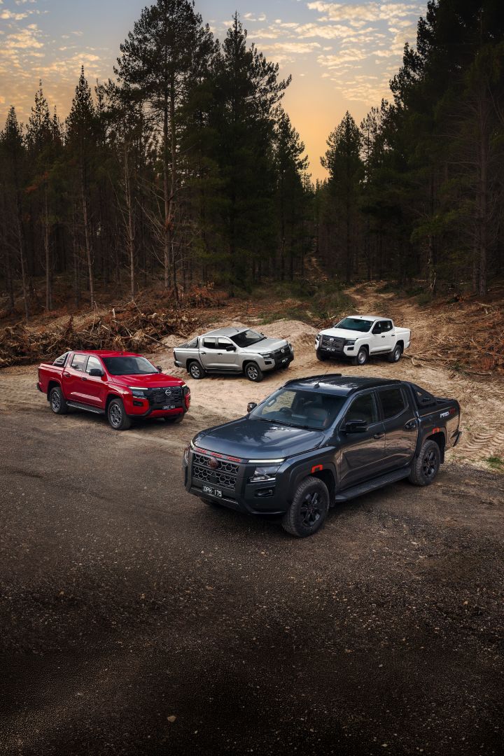 Four Nissan Navara variants parked together on a forest roadside at dusk: a Navara PRO-4X in Boulder Grey in the foreground, a Navara ST-X in Outback Red to the left, a Navara ST in Summit Silver near the centre, and a Navara SL in Alpine White to the right, with tall pine trees in the background.