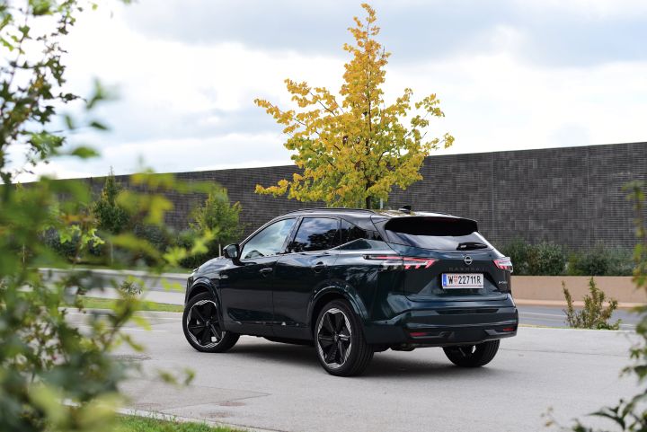 3/4 Heckansicht eines dunkelgrünen Nissan Qashqai auf einem Parkplatz neben einem schmalen Baum mit Herbstlaub
