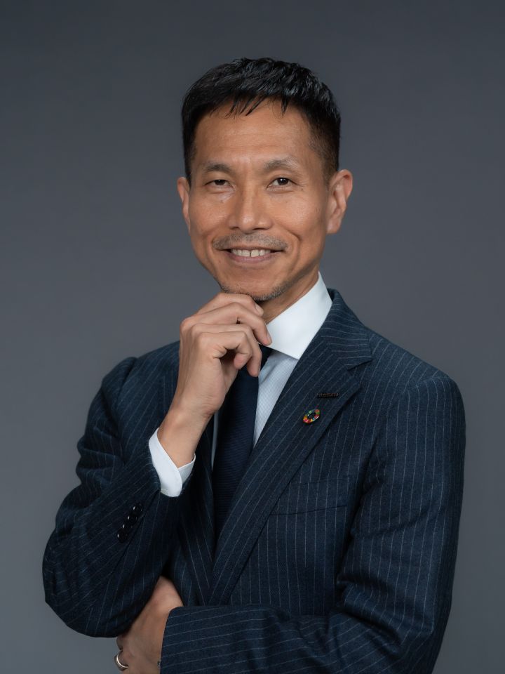 Portrait of Toru Ihara placing his hand on his chin (suit, tie, NISSAN corporate pin and SDGs badge)