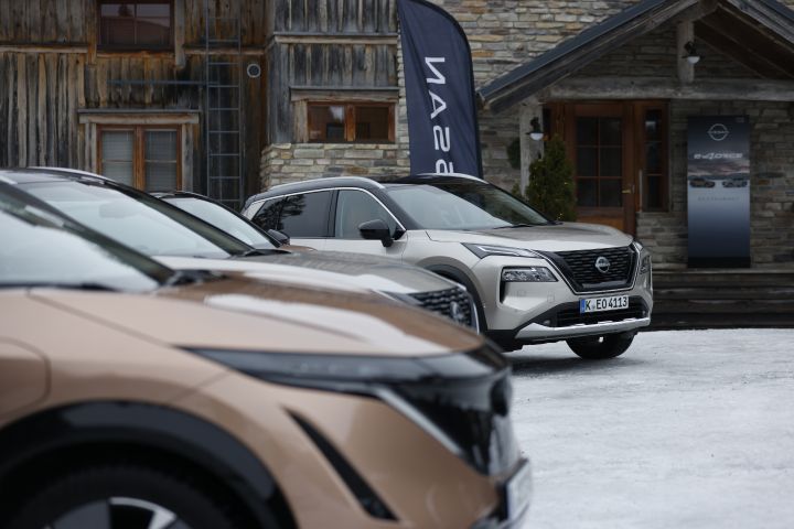 Unscharf im Vordergrund ist die Front des Nissan Elektroautos Nissan Ariya zu sehen, der Fokus liegt auf dem weiter im Hintergrund stehenden silbernen Nissan X‑Trail