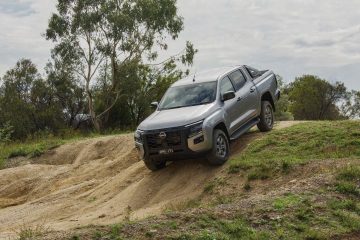 2026 Nissan Navara ST dual cab ute finished in Summit Silver shown from the rear-left on uneven off-road terrain.